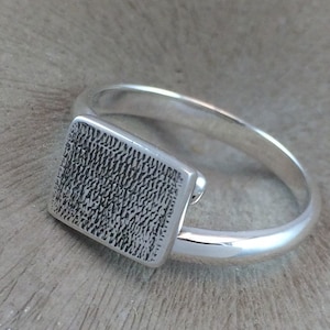 May include: A silver ring with a textured rectangular top. The ring is made of polished silver and has a simple band.