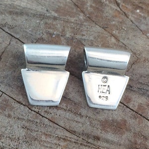 May include: Two polished silver pendant bails. Each bail has a rectangular top and a trapezoidal body. One bail is stamped with "HEA 925". The pendants are displayed on a weathered wooden surface.