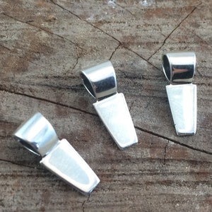 May include: Three silver-colored pendant bails on a wooden surface. Each bail has a rectangular base and a curved loop at the top. The bails are made of a shiny, reflective metal.