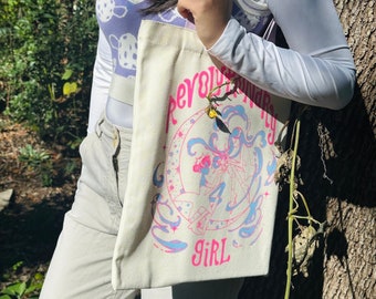 May include: A white canvas tote bag with a pink and blue design featuring a woman in a crescent moon with the text "Revolutionary Girl".