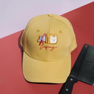 May include: Yellow baseball cap with an embroidered design of a loaf of bread with a cat face and the word "loafing" in red script. The cap is on a red surface with a black cleaver.