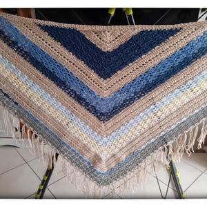 Pode incluir: Um xale triangular de crochê com um padrão em forma de V e faixas alternadas de fio bege, azul-marinho, azul-claro e creme. A peça é finalizada com longas franjas de cor creme ao longo das bordas inferiores e um ponto texturizado.
