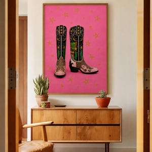 May include: A framed wall art print featuring two black cowboy boots with pink accents, leopard print details, and green cactus motifs. The boots are set against a vibrant pink background decorated with scattered gold stars throughout the composition.
