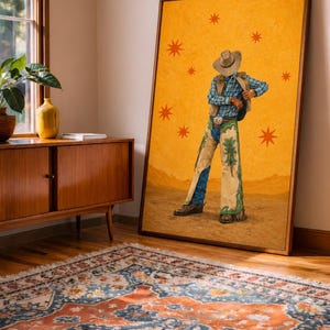 Puede incluir: Una obra de arte enmarcada que presenta una figura de vaquero con sombrero y chaparreras, sobre un fondo amarillo con motivos de estrellas. La obra está apoyada contra una pared, con una alfombra estampada y muebles de madera en primer plano.