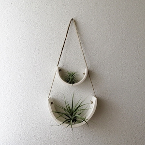 May include: Two white ceramic hanging planters with air plants. The planters are shaped like half circles and are suspended from the ceiling with twine.
