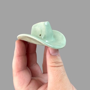 May include: A small, light green ceramic cowboy hat with a hole in the top.