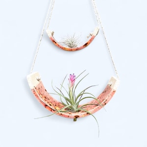 May include: Two-tiered hanging planter with a horseshoe design. The planters are made of a reddish-orange material with a speckled pattern and are suspended by white rope. The lower planter holds a flowering plant with a pink bloom.