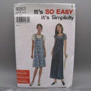 Könnte beinhalten: Ein Simplicity-Schnittmusterumschlag 9263, Größe A 8-18, mit Abbildungen von zwei Latzkleidern. Eines ist ein blau kariertes Kleid mit Blumenmuster, das andere ein dunkelblaues Kleid im Jeans-Stil, beide über T-Shirts getragen.