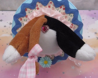 May include: A plush toy head featuring one brown ear and one black ear, both with white tips. The toy has a pink ruffled collar, a pink heart-shaped gem, and a light pink gingham ribbon tied to the brown ear. It is mounted on a circular base.