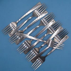 May include: An assortment of stainless steel forks on a blue background. Each fork features a slender handle and four tines. The forks are marked with the word "DELTA" and a decorative arrow design.
