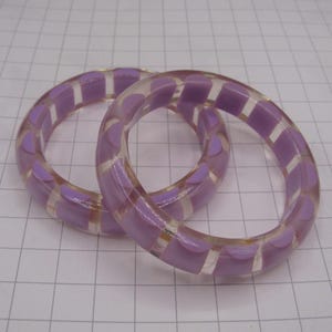 May include: Two clear bangles with a lavender and white geometric design. The bracelets are made of a transparent material and feature a repeating pattern of lavender shapes separated by clear sections. The bangles are circular in shape.