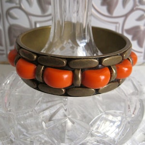 May include: A vintage brass bangle bracelet with orange resin accents. The bracelet features a series of orange, half-circle shaped beads set within a dark metal frame. The bracelet is displayed on a clear glass object.
