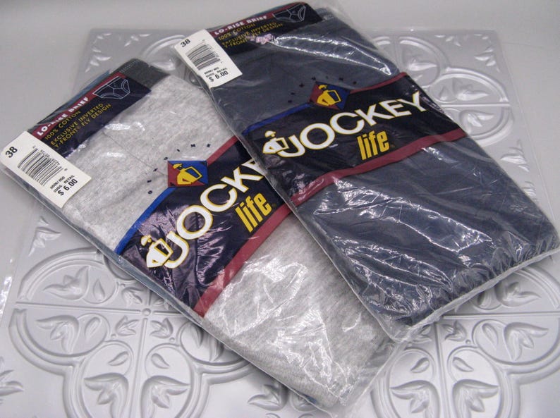 May include: Two packages of Jockey Life brand briefs, one in heather gray and one in navy blue. Each package is sealed in clear plastic and features a dark blue label with the Jockey Life logo, size 38, and text indicating 100% cotton material.
