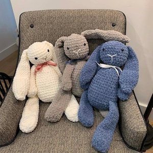 May include: Three handmade crochet bunny plushies in white, gray, and blue. Each bunny has long floppy ears, a stitched face, and a ribbon bow. The bunnies are sitting on a gray upholstered chair.