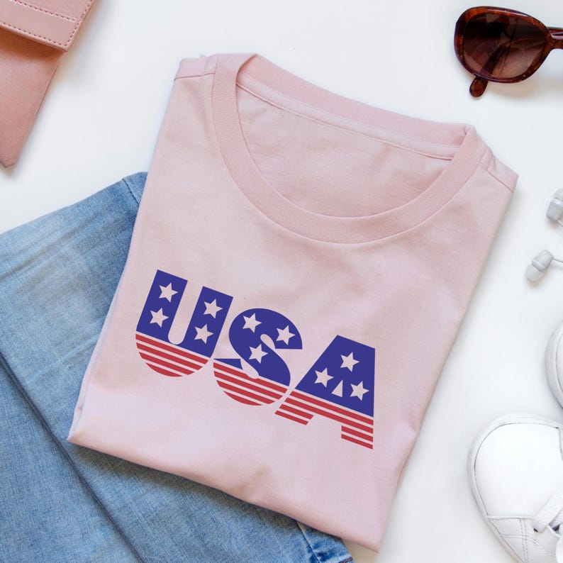May include: A light pink t-shirt with the word "USA" printed in blue and red. The letters are designed with a star pattern and stripes, evoking the American flag. Sunglasses, a wallet, and sneakers are also visible.
