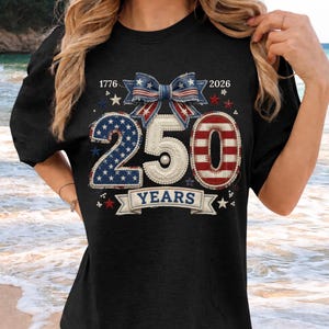 Peut inclure: T-shirt noir avec un motif patriotique célébrant 250 ans. Le motif comprend les chiffres "250" en rouge, blanc et bleu, un nœud, des étoiles et les années "1776" et "2026". Le mot "YEARS" est affiché sur une bannière.