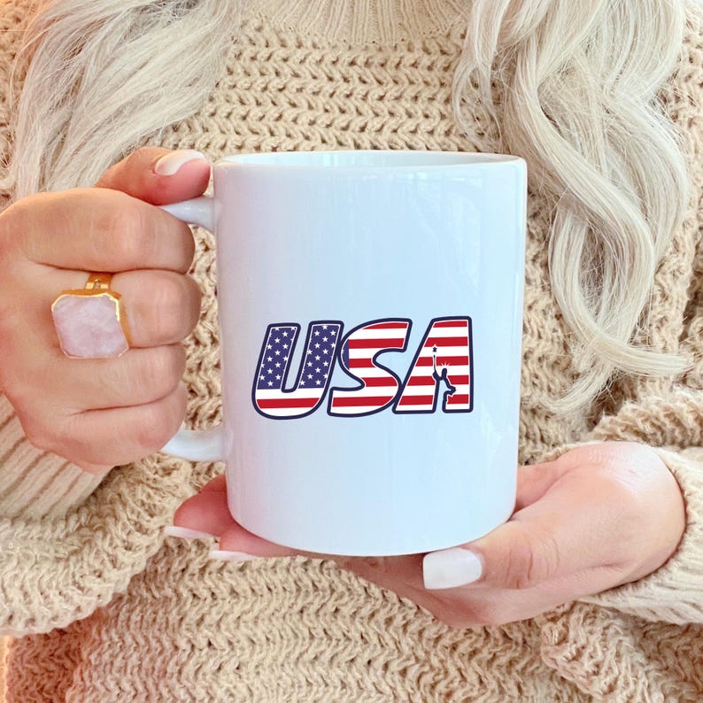 May include: White ceramic mug featuring the word "USA" in red, white, and blue, with a design reminiscent of the American flag. The mug has a standard handle and a smooth, glossy finish.