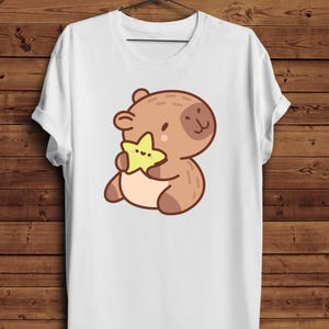 May include: White t-shirt featuring a cartoon bear holding a yellow star. The bear is brown with darker brown accents and a small, black dot for an eye. The star is yellow with three small dots. The shirt is hanging on a wooden background.