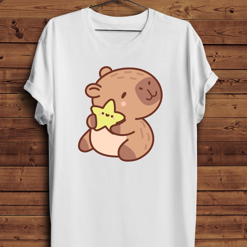 May include: White t-shirt featuring a cartoon bear holding a yellow star. The bear is brown with darker brown accents and a small, black dot for an eye. The star is yellow with three small dots. The shirt is hanging on a wooden background.