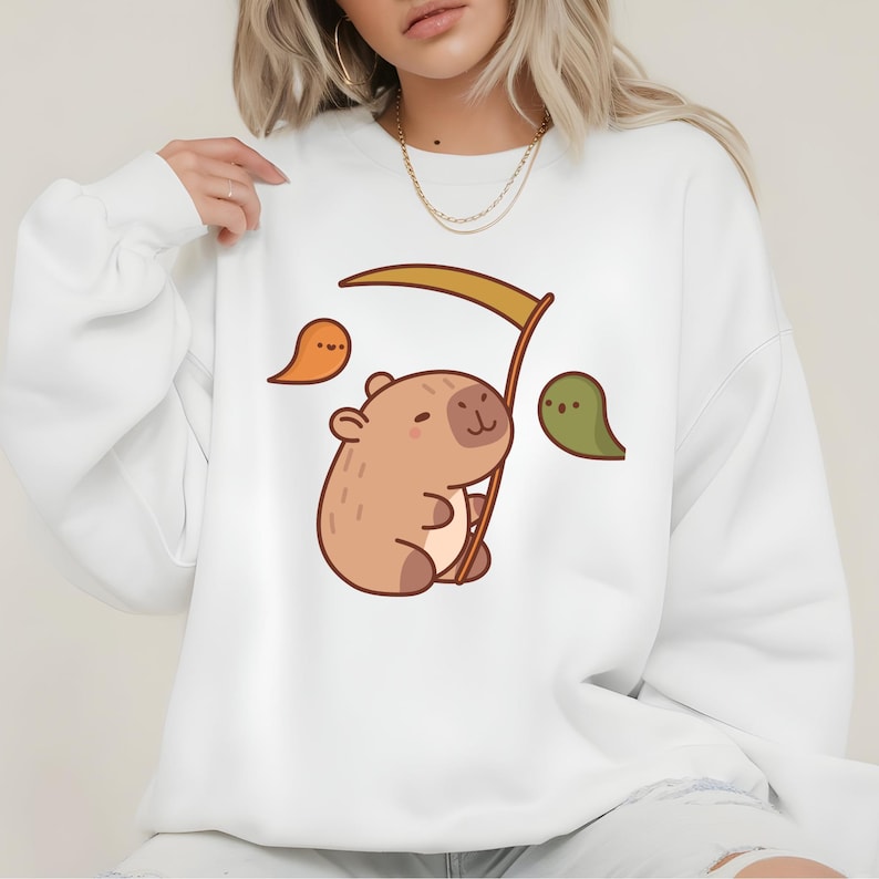 May include: White sweatshirt featuring a cartoon capybara holding a scythe. The capybara is light brown with a smiling face. Two cartoon ghosts in orange and green are on either side of the capybara.