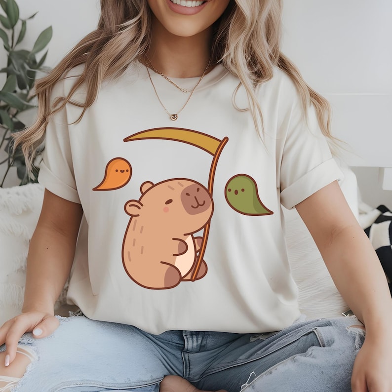 May include: Cream-colored t-shirt featuring a cartoon capybara holding a scythe, with two ghost-like figures on either side. The capybara is brown and tan, and the ghosts are orange and green. The shirt is made of a soft material.
