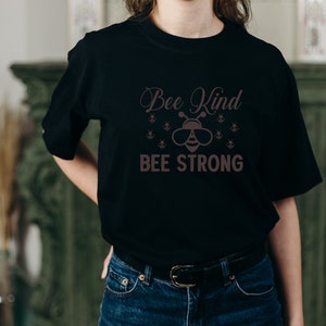 Może przedstawiać: Czarna koszulka z napisem "Bee Kind BEE STRONG" w brązowej czcionce. Projekt zawiera ilustrację pszczoły w okularach przeciwsłonecznych i kilka mniejszych grafik pszcz&oacute;ł.