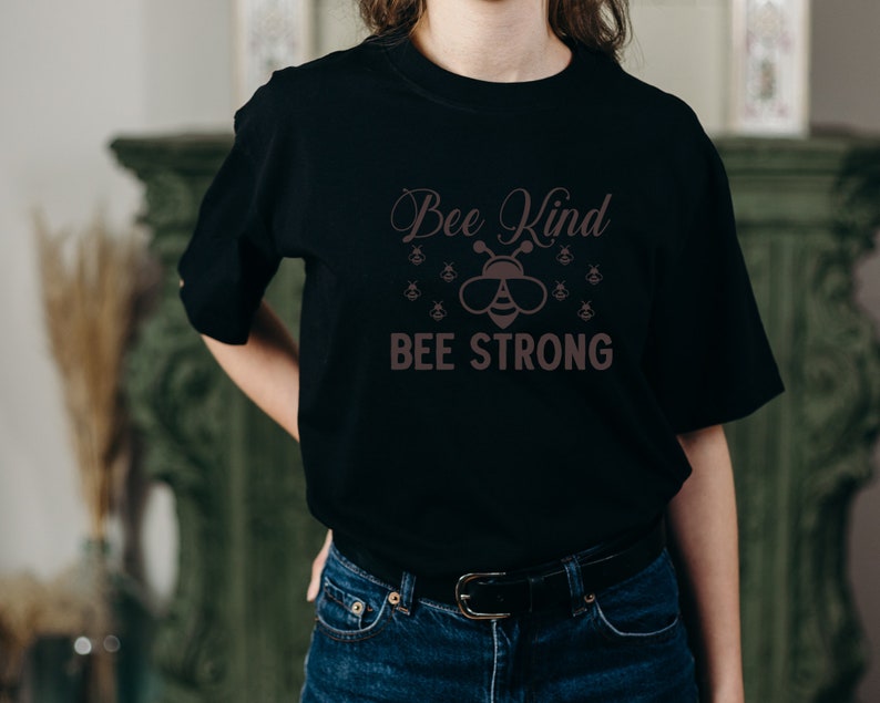 Może przedstawiać: Czarna koszulka z napisem "Bee Kind BEE STRONG" w brązowej czcionce. Projekt zawiera ilustrację pszczoły w okularach przeciwsłonecznych i kilka mniejszych grafik pszcz&oacute;ł.