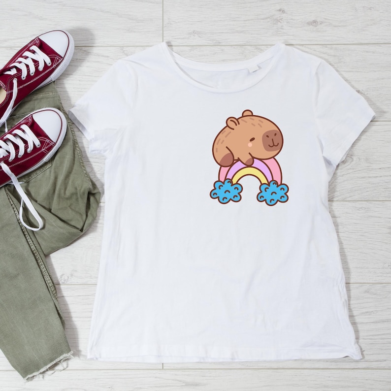 May include: White t-shirt featuring a cartoon capybara resting on a rainbow. The capybara is brown, and the rainbow has pastel colors. The design includes blue clouds at the rainbow's ends. Casual wear.