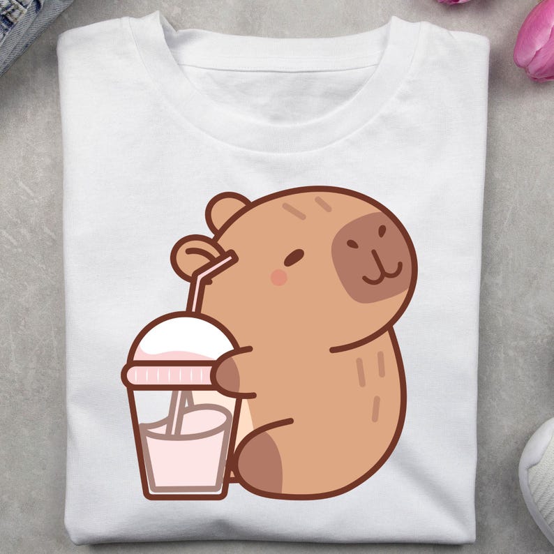 May include: White t-shirt featuring a cartoon capybara holding a pink drink with a straw. The capybara is light brown with a small, smiling face. The drink is in a clear cup with a white lid and pink accents.