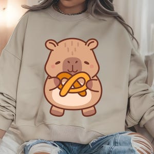 May include: Beige sweatshirt featuring a cartoon bear holding a pretzel. The bear is brown with a light brown belly and a pretzel in its paws. The sweatshirt is a solid beige color.