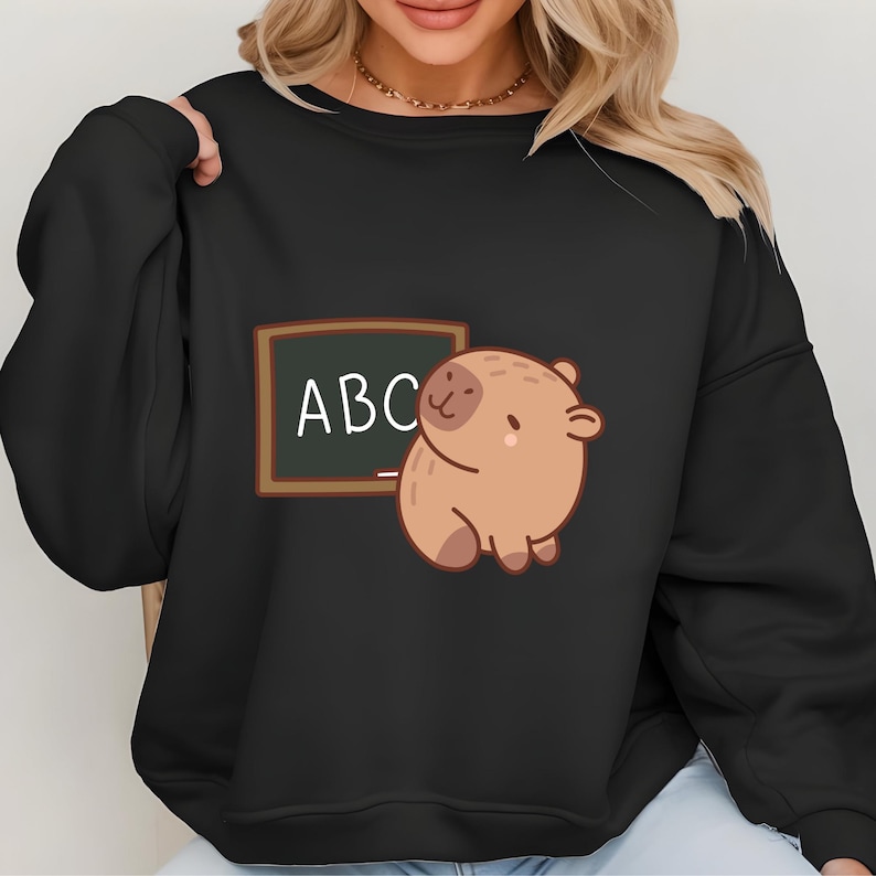 May include: Black sweatshirt featuring a cartoon capybara next to a chalkboard with the letters "ABC" in white. The capybara is light brown with a small nose and feet. The chalkboard has a brown frame.