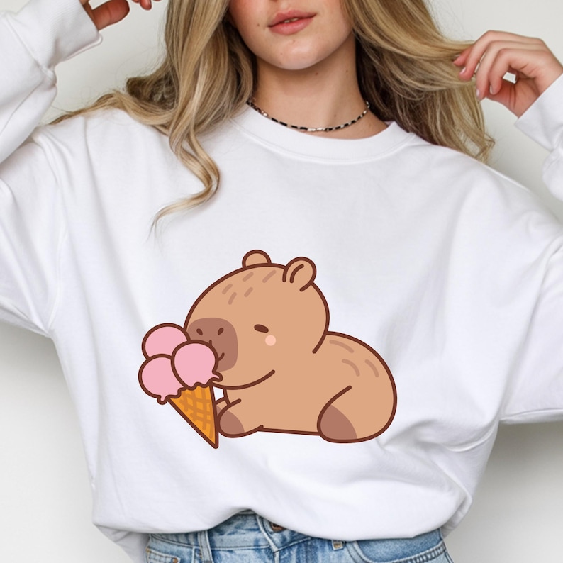 May include: White sweatshirt featuring a cartoon capybara enjoying a pink ice cream cone. The capybara is light brown with a small, smiling face. The ice cream cone is a light brown color.