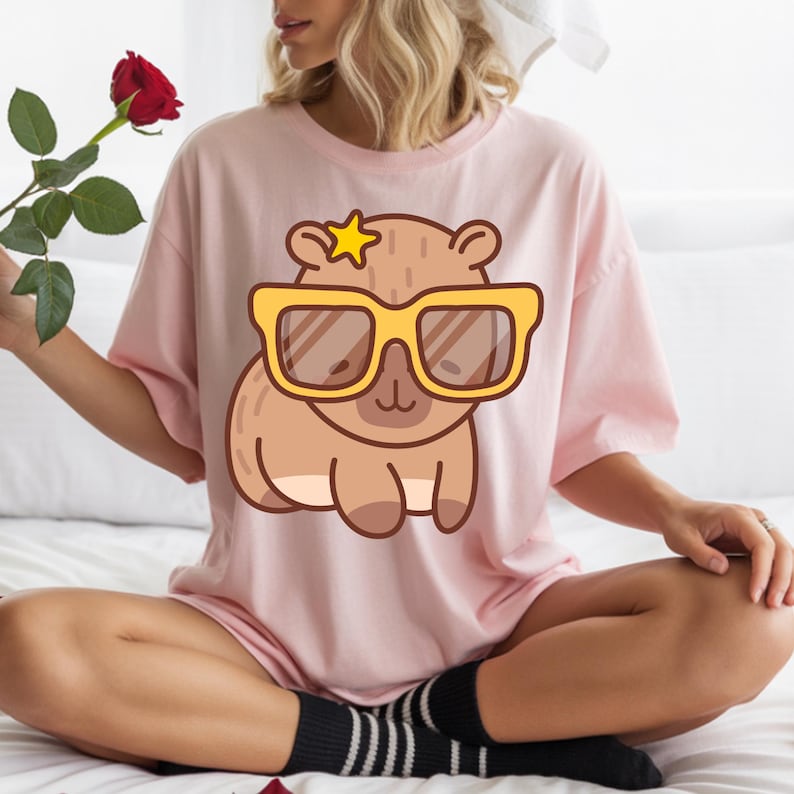 May include: A light pink t-shirt featuring a cartoon capybara wearing yellow sunglasses and a star. The capybara is brown with a light brown belly. The shirt is a relaxed fit.