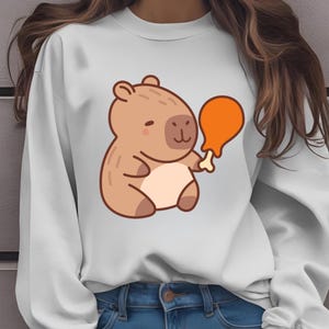 May include: White sweatshirt featuring a cartoon capybara holding a drumstick. The capybara is brown with a light beige belly. The drumstick is orange. The sweatshirt is a solid white color.