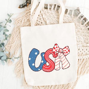 May include: Cream-colored tote bag with the word "USA" in red, white, and blue. The "U" is blue with white stars, the "S" is red with white stripes, and the "A" has a red and white checkered bow.