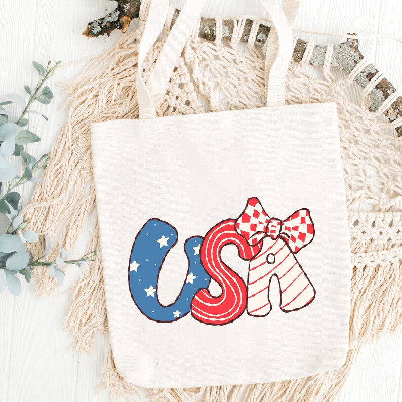 May include: Cream-colored tote bag with the word "USA" in red, white, and blue. The "U" is blue with white stars, the "S" is red with white stripes, and the "A" has a red and white checkered bow.