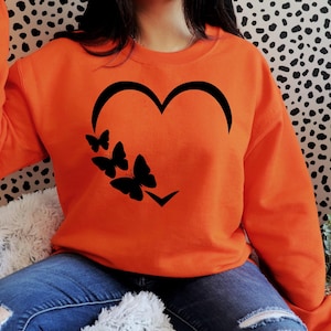 Pu&ograve; includere: Felpa arancione con un design a forma di cuore nero e quattro farfalle nere all'interno del cuore.
