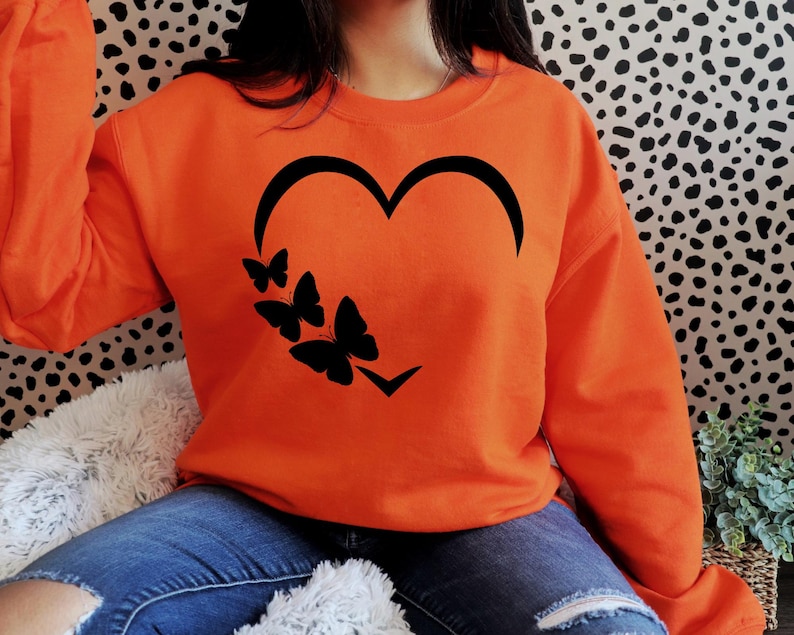 Pu&ograve; includere: Felpa arancione con un design a forma di cuore nero e quattro farfalle nere all'interno del cuore.