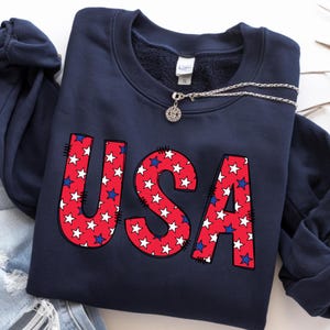 May include: Navy blue sweatshirt with the word "USA" in red letters filled with white stars and blue stars. A silver necklace with a pendant is draped across the sweatshirt. The sweatshirt is folded on top of a pair of blue jeans.