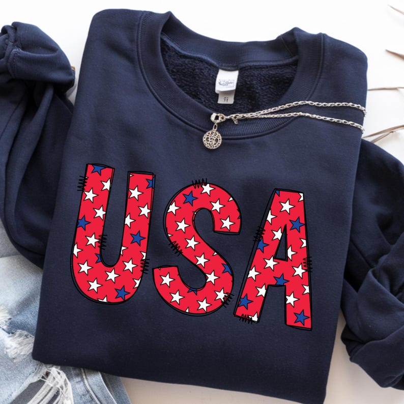 May include: Navy blue sweatshirt with the word "USA" in red letters filled with white stars and blue stars. A silver necklace with a pendant is draped across the sweatshirt. The sweatshirt is folded on top of a pair of blue jeans.