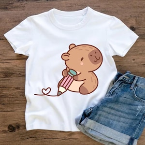 May include: White t-shirt featuring a cartoon bear holding a pink and gray pencil, with a heart-shaped line extending from the pencil tip. The bear is brown with a light tan belly. Denim shorts are visible in the image.