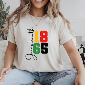 May include: A cream-coloured t-shirt with the word "Juneteenth" in black script, and the numbers "1865" in yellow, red, green, and black. The t-shirt is a casual style.