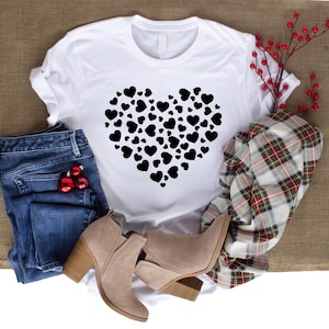 Pu&ograve; includere: Maglietta bianca con un design a forma di cuore nero composto da cuori pi&ugrave; piccoli. La maglietta &egrave; appoggiata su una superficie di juta marrone con un paio di jeans blu, una sciarpa a quadri e due stivaletti marroni.