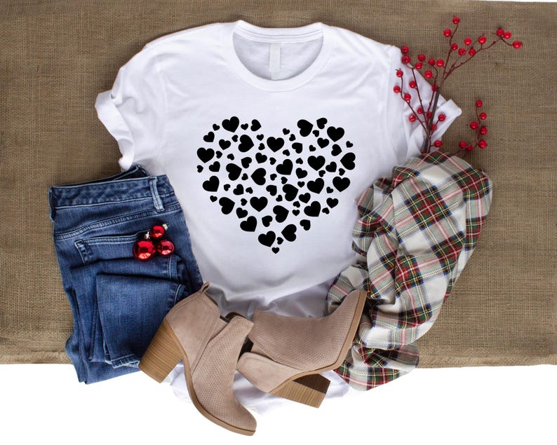 Pu&ograve; includere: Maglietta bianca con un design a forma di cuore nero composto da cuori pi&ugrave; piccoli. La maglietta &egrave; appoggiata su una superficie di juta marrone con un paio di jeans blu, una sciarpa a quadri e due stivaletti marroni.