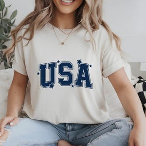May include: Cream-colored t-shirt with the word "USA" in navy blue block letters, accented with small stars. The shirt has short sleeves and is made of a soft material. A casual and patriotic design.