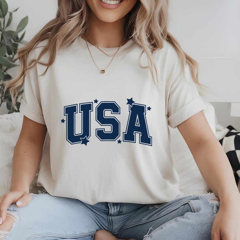 May include: Cream-colored t-shirt with the word "USA" in navy blue block letters, accented with small stars. The shirt has short sleeves and is made of a soft material. A casual and patriotic design.