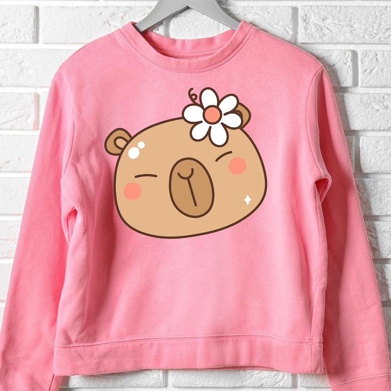 May include: Pink sweatshirt featuring a cartoon bear face with a white flower on its head. The bear has closed eyes, rosy cheeks, and a diamond-shaped sparkle. The sweatshirt is hanging on a gray hanger against a white brick wall.