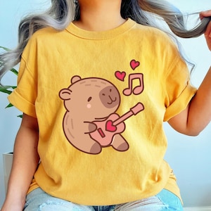 May include: Mustard yellow t-shirt featuring a cartoon bear playing a pink guitar with a heart-shaped sound hole. Two pink hearts and a musical note float above the bear. The shirt has rolled-up sleeves.