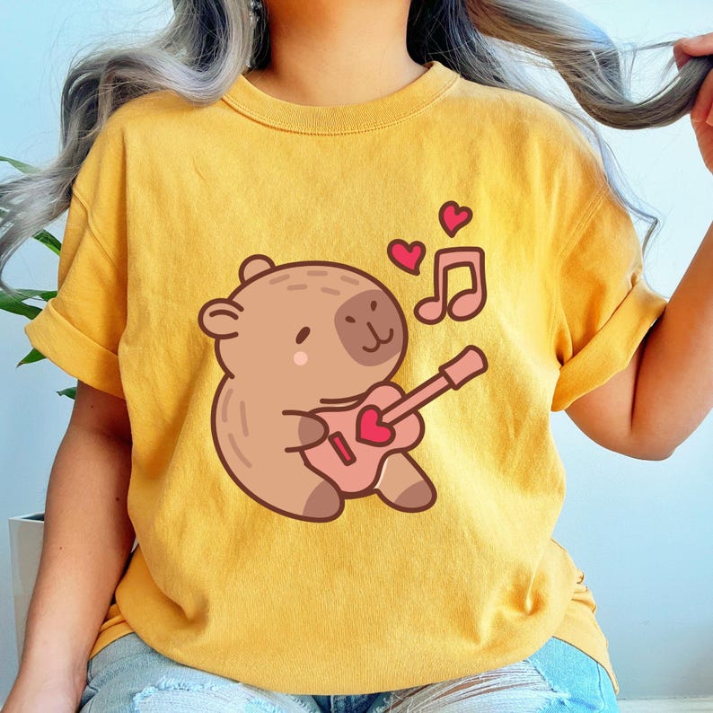 May include: Mustard yellow t-shirt featuring a cartoon bear playing a pink guitar with a heart-shaped sound hole. Two pink hearts and a musical note float above the bear. The shirt has rolled-up sleeves.