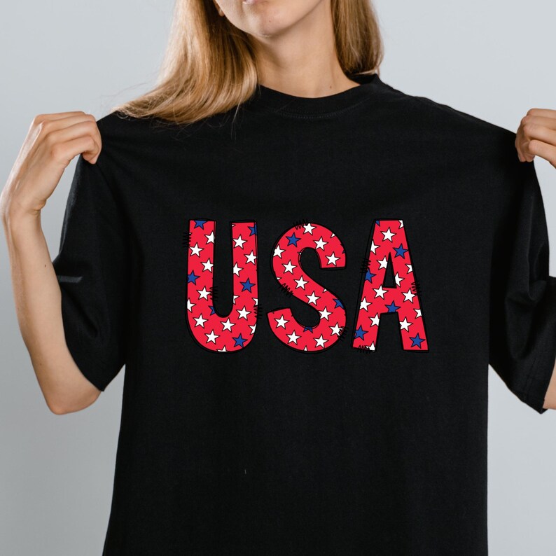 May include: Black t-shirt with the word "USA" in large, bold letters. The letters are red with a white star pattern, and outlined in blue. The shirt is a casual style.